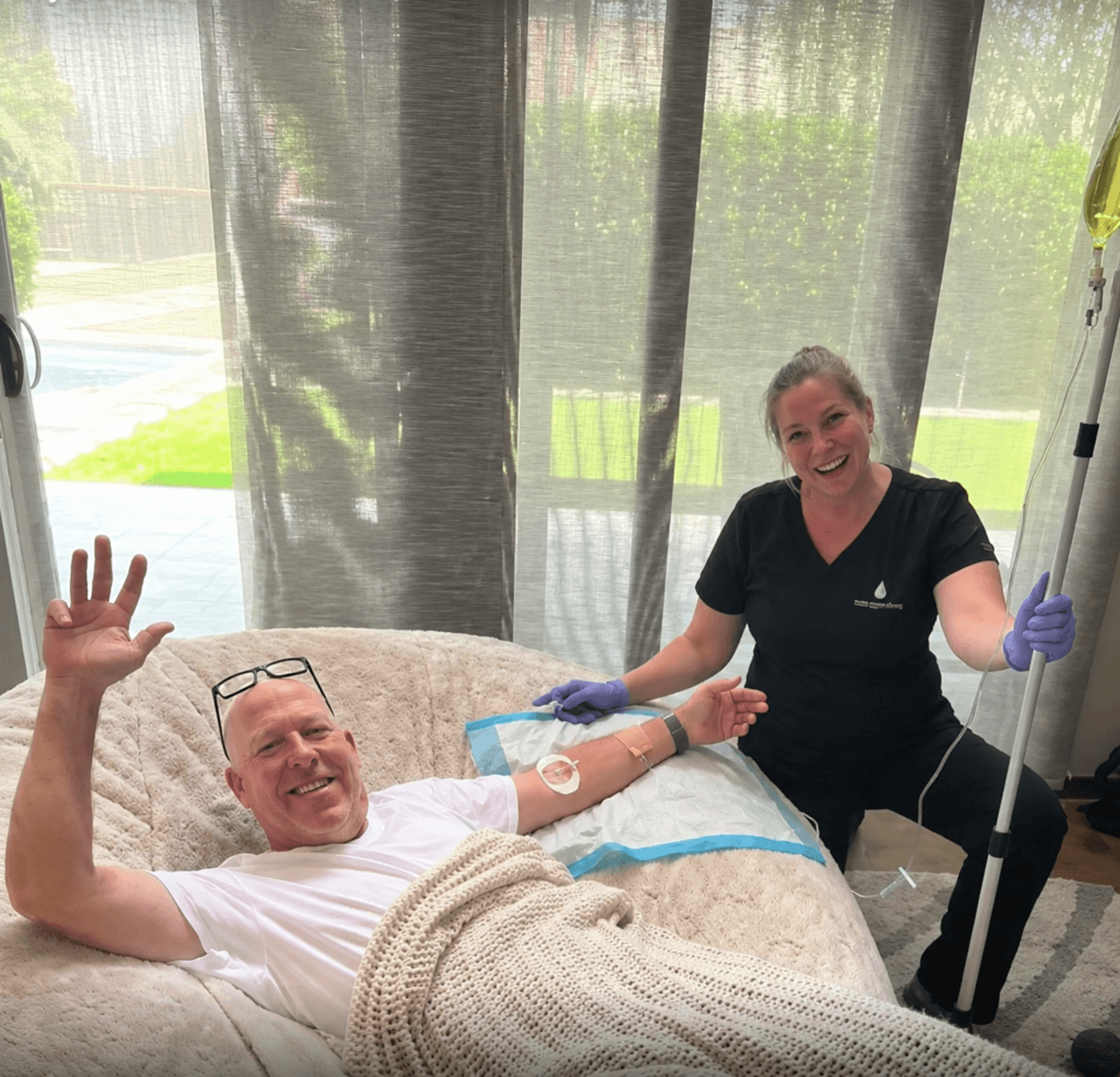A relaxed client waving while receiving an in-home IV drip from a smiling Prime IV licensed medical professional in Mesa, AZ