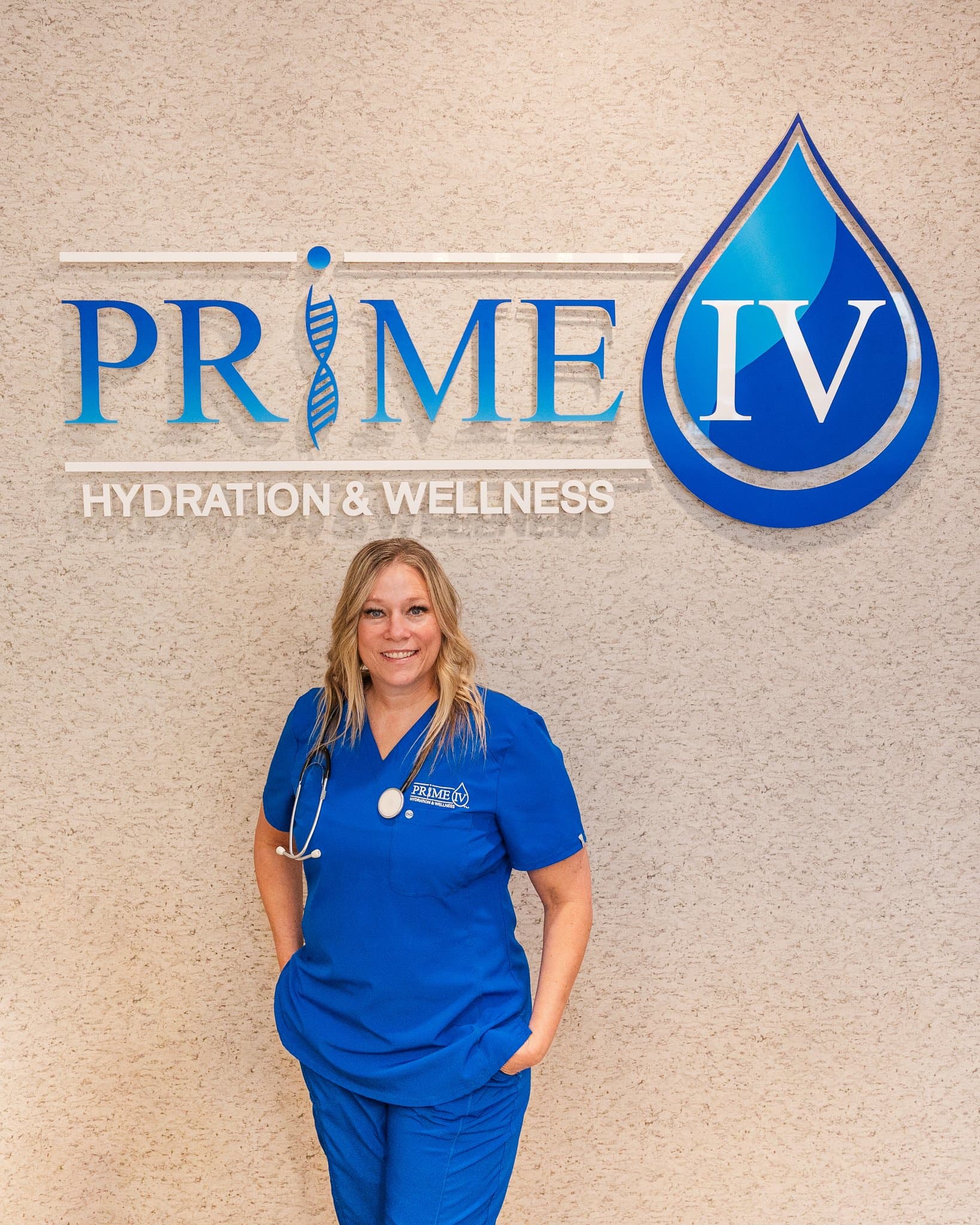 Nurse Tiffany, Lead Registered Nurse at Prime IV Mesa, in Prime IV scrubs with a stethoscope in front of the Prime IV Hydration & Wellness logo wall